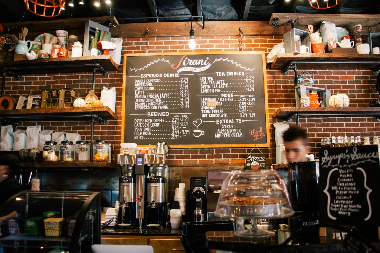 A barista at work behind a coffee counter, beside a chalkboard listing syrups and bottles on the shelf