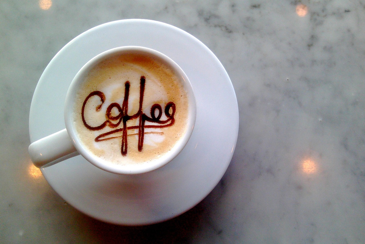 A cappuccino with the word Coffee drawn in chocolate on the foam, in a white cup and saucer on a marble table